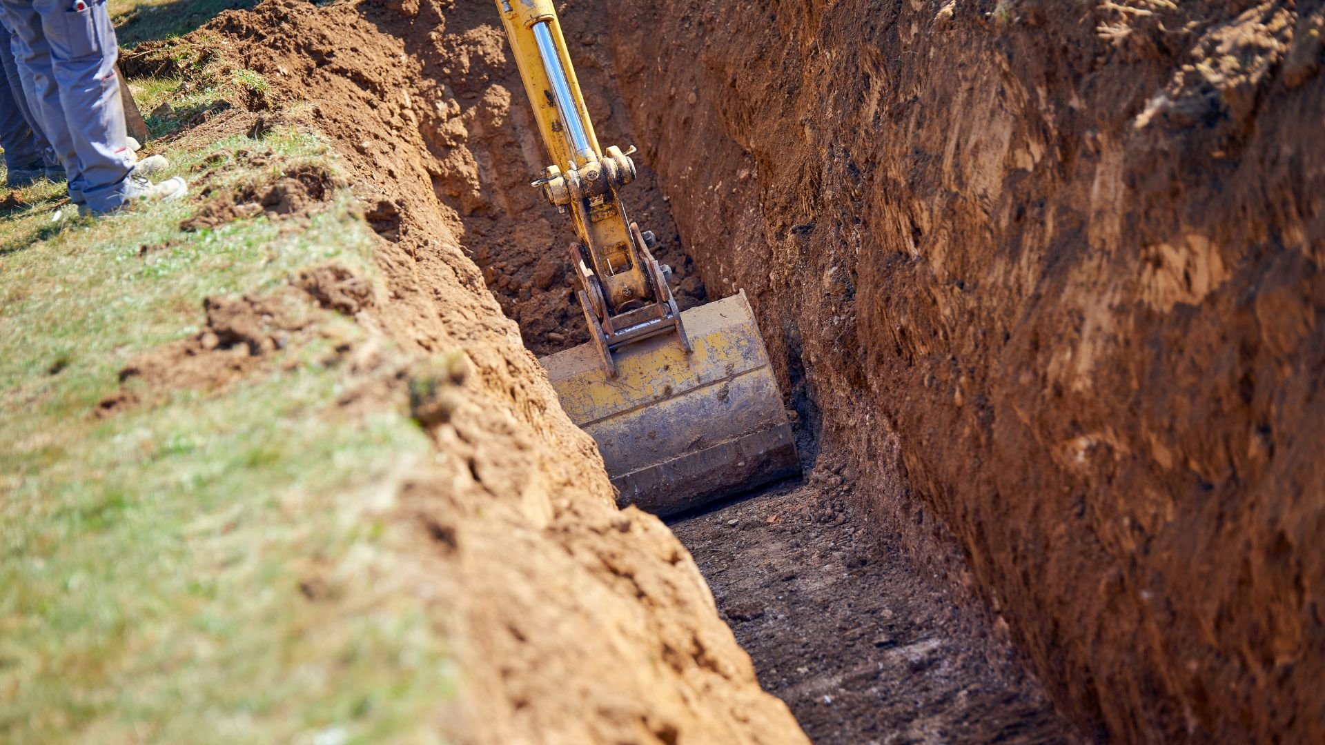 Excavator digging deep trench in dirt with grass and soil mounds