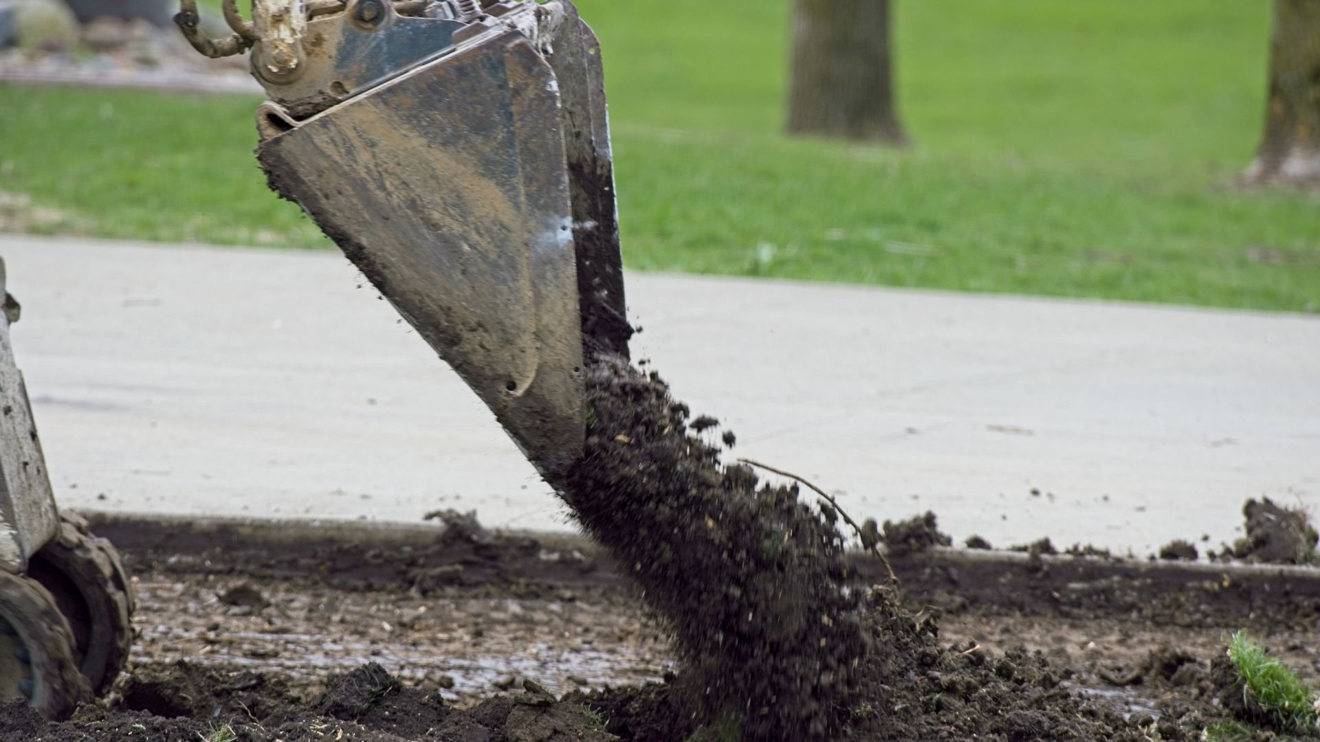 Excavator bucket digging and moving dirt near concrete sidewalk and grass