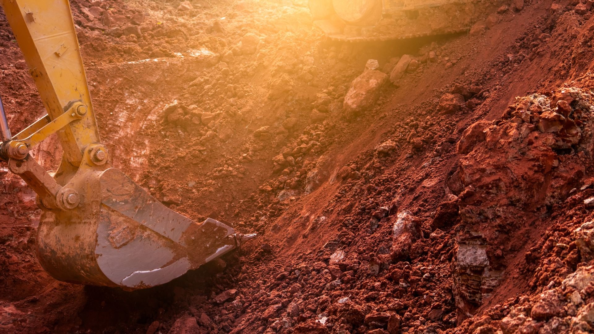 Excavator bucket digging into red rocky soil under warm sunlight